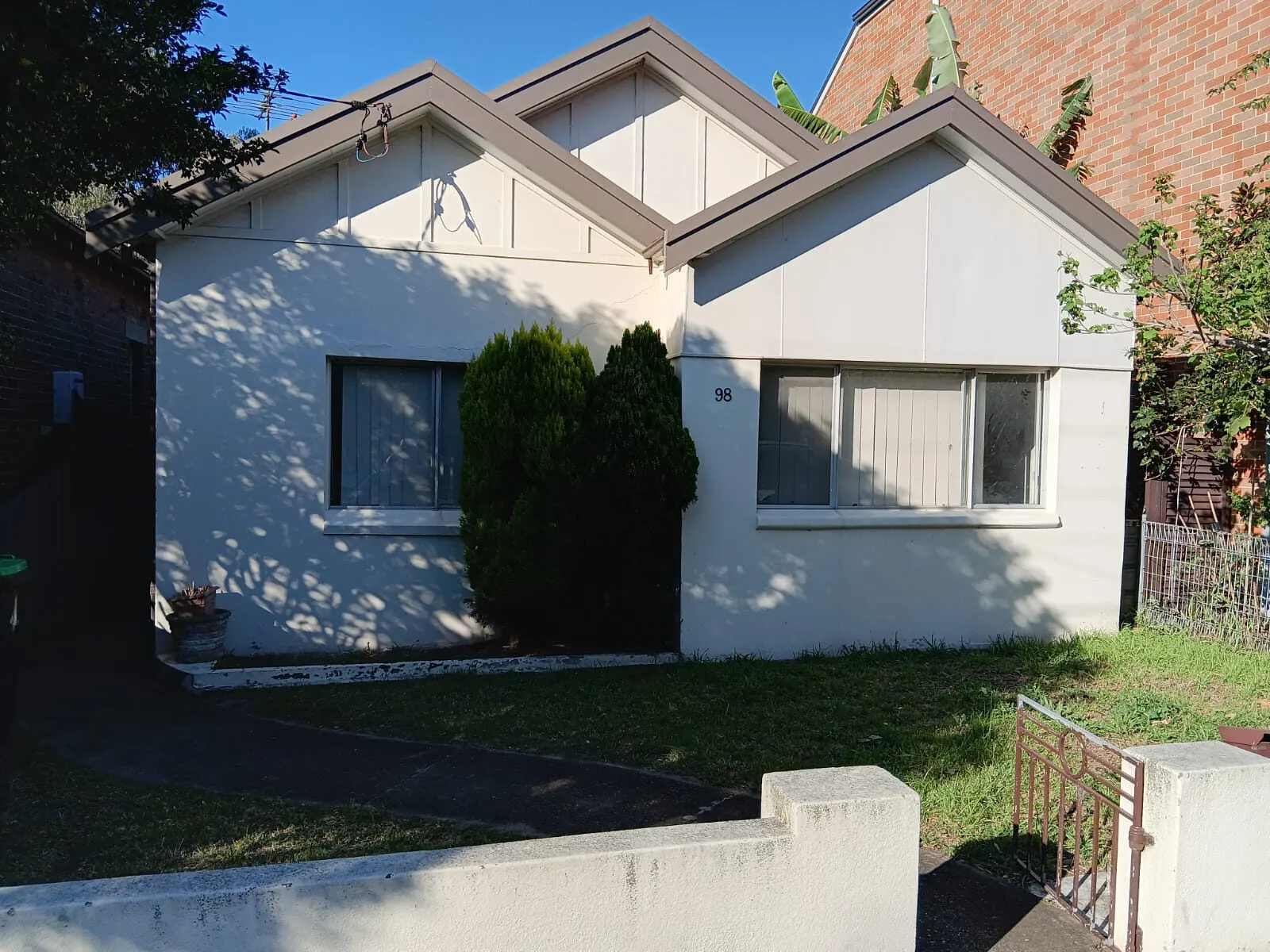 A white rendered cottage in Mascot with a small front garden and a shaded front lawn.